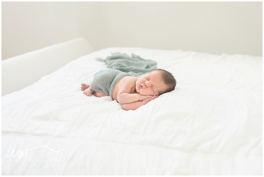 Newborn baby boy laying on an all white bed sleeping with his hands under his head. 
