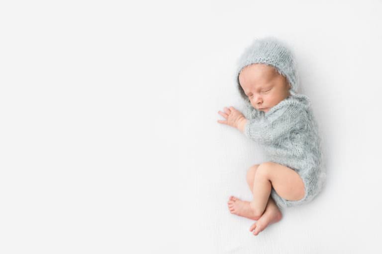 a tiny baby in a light blue onesie and bonnet sleeping on a white bed