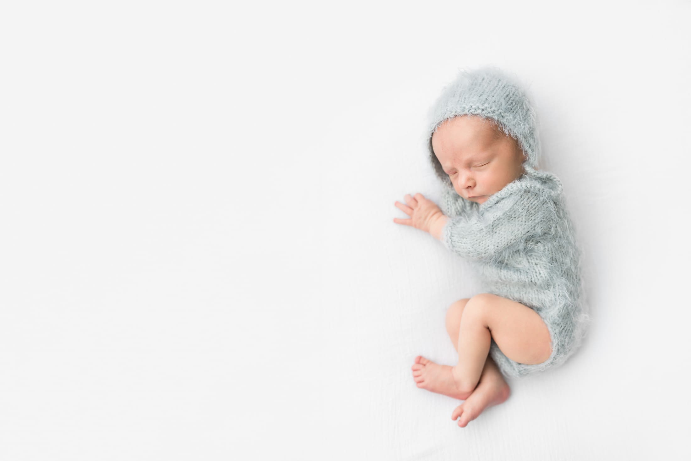 a tiny baby in a light blue onesie and bonnet sleeping on a white bed